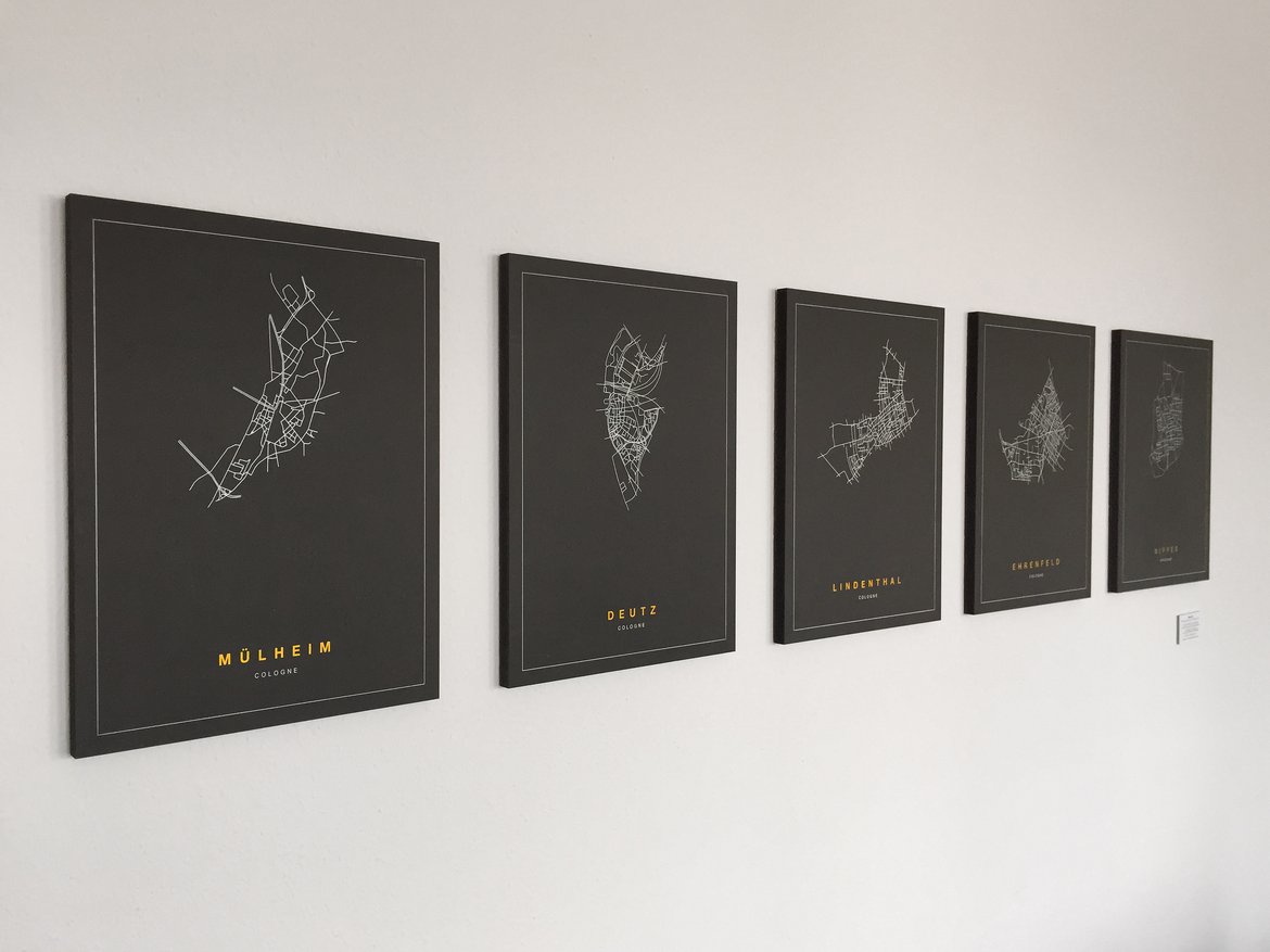 Mehrere Bilder mit schwarzem Hintergrund an einer weißen Wand. Abgebildet sind Stadteile Kölns mit weißen Linien