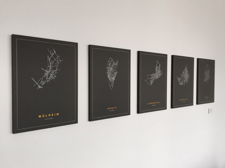 Mehrere Bilder mit schwarzem Hintergrund an einer weißen Wand. Abgebildet sind Stadteile Kölns mit weißen Linien